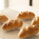 Pãezinhos tipo pretzel artesanais com cortes no topo e sal grosso esfriando sobre grade de metal.