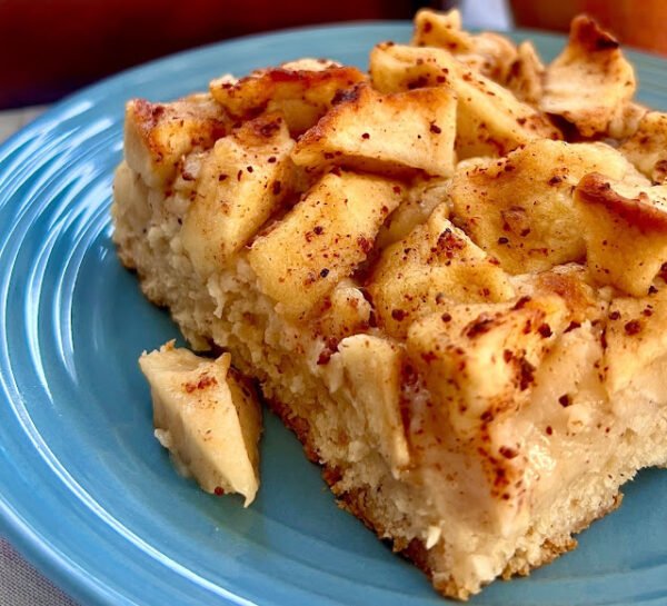 Fatia de bolo de maçã com canela fofinho servida em um prato azul com pedaços da fruta por cima.