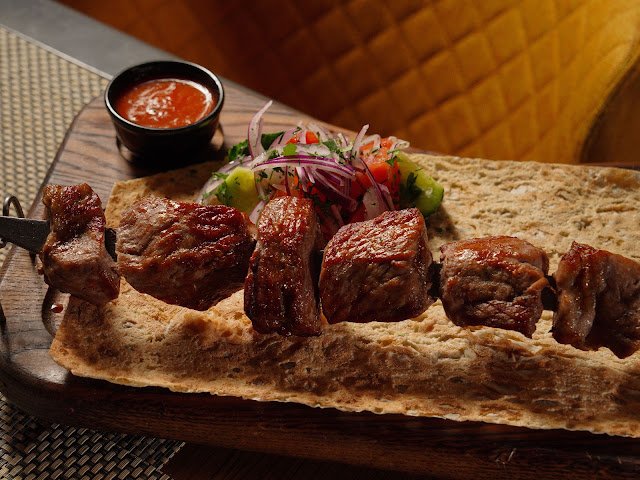 Espetinho de carne bovina grelhada e suculenta servido sobre pão folha em uma tábua de madeira, acompanhado de vinagrete de cebola roxa e molho vermelho.