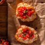Dois molletes mexicanos dispostos sobre papel pardo. O pão francês cortado ao meio está tostado, coberto com feijão refrito, queijo muçarela derretido e finalizado com vinagrete pico de gallo colorido por cima.