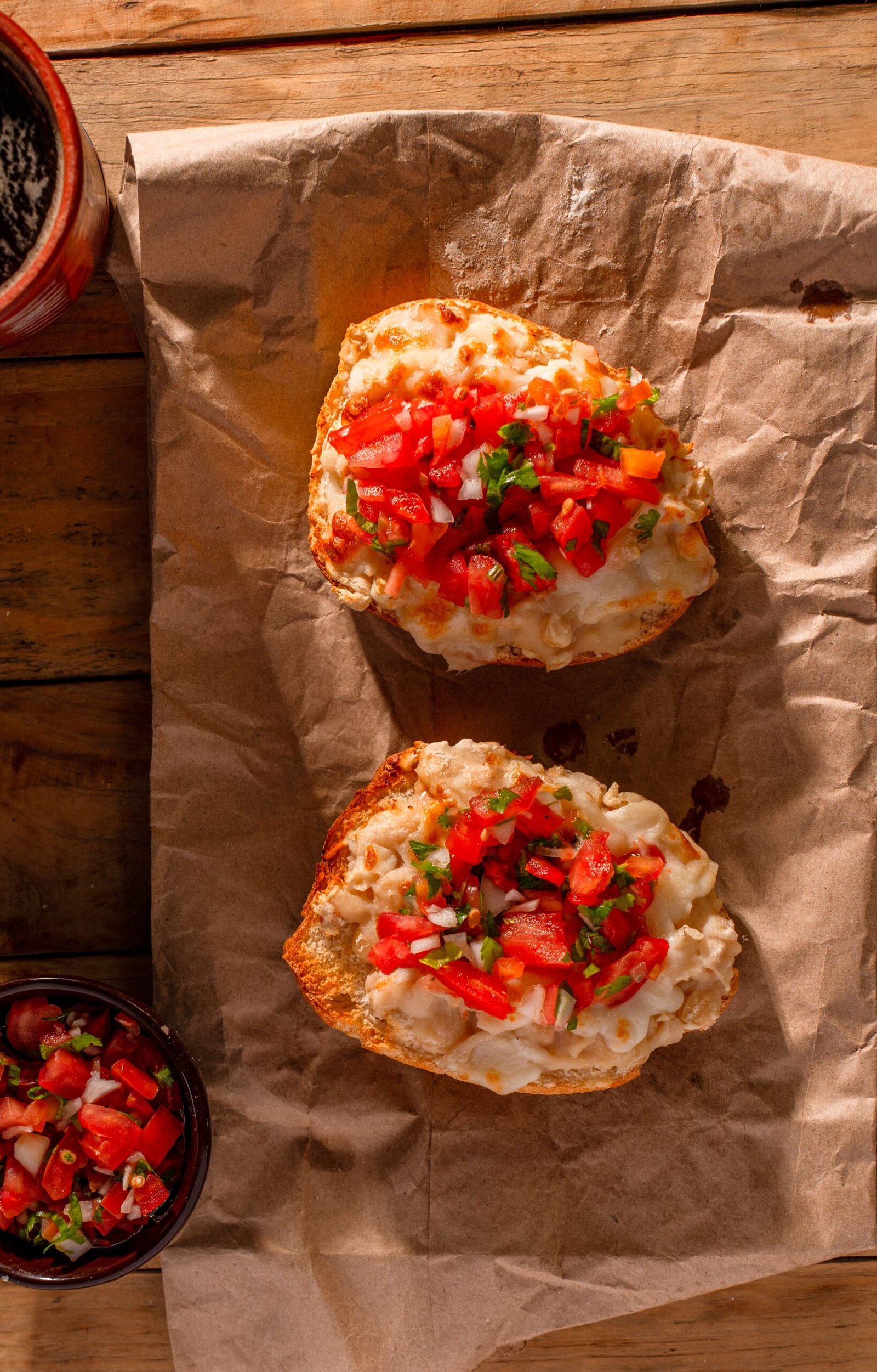 Dois molletes mexicanos dispostos sobre papel pardo. O pão francês cortado ao meio está tostado, coberto com feijão refrito, queijo muçarela derretido e finalizado com vinagrete pico de gallo colorido por cima.