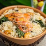 Tigela rústica de barro cheia de arroz paraense cremoso com tucupi, folhas verdes de jambu e camarões grelhados inteiros no topo.